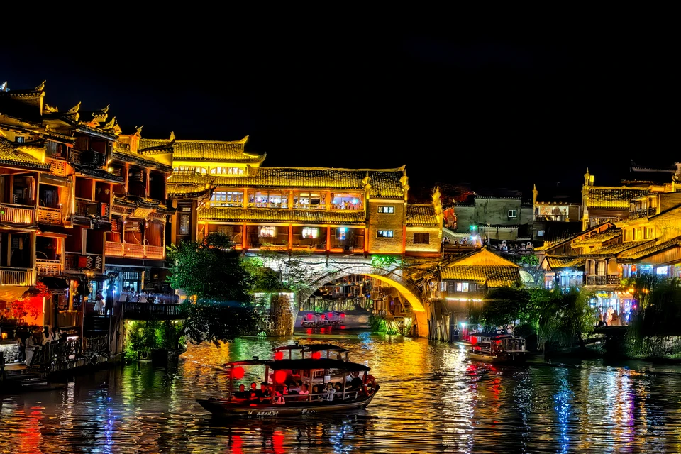 Lantern-lit riverside night view of Fenghuang Ancient Town with traditional bridges and reflections.
